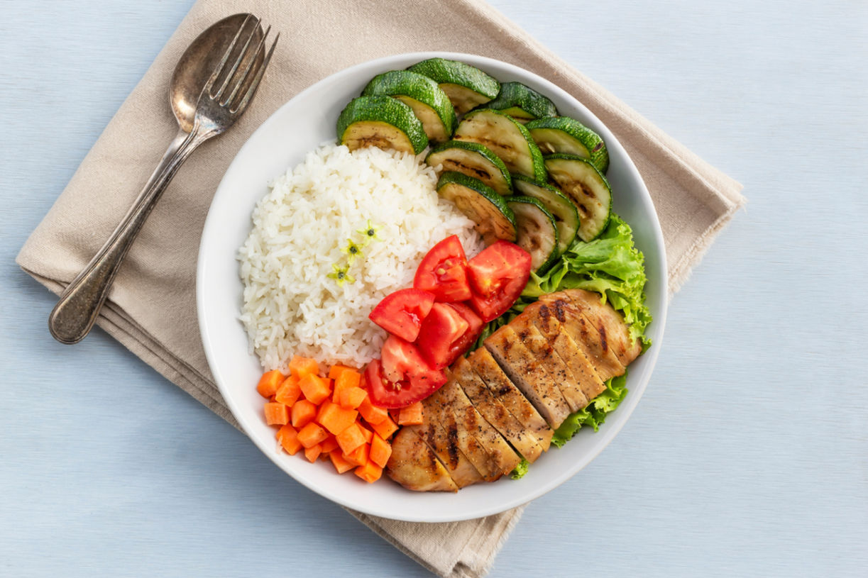 The image shows a plate with a balanced and colorful meal. It contains white rice, grilled chicken breast, sautéed zucchini slices, diced tomatoes, cubed carrots, and a side of green lettuce leaves. A fork is placed next to the plate on a beige cloth napkin or placemat against a light blue background